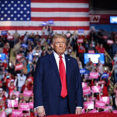 Donald Trump stands in front of a large crowd at a political rally, with a giant American flag hanging behind him. He is wearing a navy blue suit, white shirt, and red tie, looking forward with a serious expression. The crowd in the background holds up signs in red, white, blue, and pink.