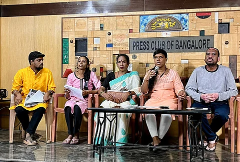Five people sit at a table on a stage with a "PRESS CLUB OF BANGALORE" backdrop. Individuals in a yellow kurta, pink top, sari, peach top speaking into a microphone, and a grey shirt are visible.