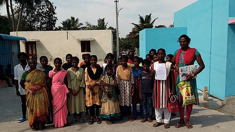 A group of Irula men, women, and children stand together outside a building in Kunnapattu village. One woman holds up a petition paper, symbolising their collective demand for land rights and housing.