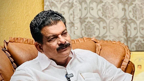 PV Anvar, seated on a brown leather chair, wearing a white shirt and mundu, engaged in a conversation, with a microphone clipped to his shirt