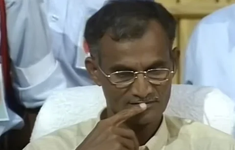 A middle-aged man wearing glasses is seated, appearing deep in thought with his finger resting against his lips. He has short hair and a mustache, and is dressed in a light-colored shirt. The background shows other people in uniforms, suggesting a formal or official setting.