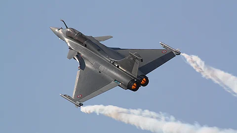 A Dassault Rafale fighter jet is seen in flight during an aerial maneuver, with both engines glowing in afterburner mode and white vapor trails streaming from its wingtips against a clear blue sky. The aircraft bears French Air Force roundels.
