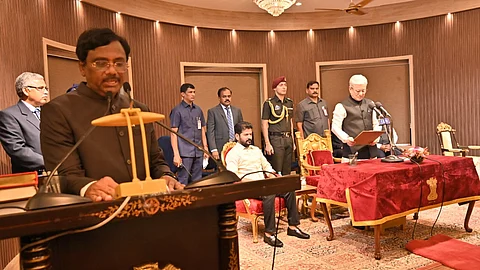 A formal swearing-in ceremony is taking place in an ornate hall with a chandelier and wooden paneling. A man in a black suit speaks at a podium with the Indian national emblem. In the background, another man in a cream-colored vest stands at a microphone reading from a document, taking the oath. Several officials, including security personnel and dignitaries, are present, some seated on red and gold chairs. A table with a red velvet cover bearing the Indian emblem is in front of the man taking the oath.