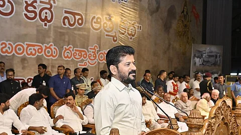 Telangana Chief Minister A. Revanth Reddy speaks at a podium during a public event, surrounded by seated dignitaries and attendees. The background features a large Telugu-language banner and historical imagery. The ornate wooden podium and richly colored floor tiles add to the ceremonial atmosphere.