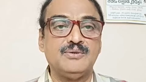 A middle-aged man with short black hair, a trimmed moustache, and large maroon-rimmed glasses is seen speaking. He is wearing a multicolored checkered shirt and is positioned against a plain indoor background with a Telugu signboard visible on the wall behind him.
