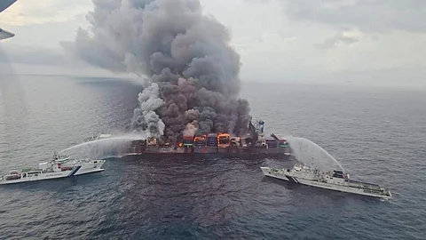 Aerial view of a large container ship engulfed in thick black smoke and flames, with two Coast Guard vessels spraying water to extinguish the fire at sea