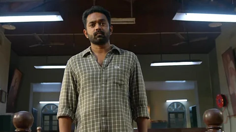 Asif Ali in the film, facing the camera, wearing a chequered shirt and standing in a courtroom.