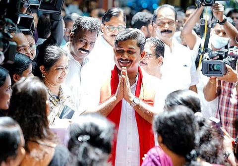 M Swaraj greets a crowd with folded hands during a campaign event, surrounded by supporters and media personnel.