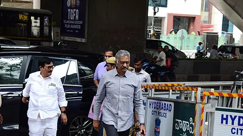 Two men, including a senior official in a grey shirt and dark trousers, are walking past police barricades marked "Hyderabad City Police" near the Jubilee Hills Law & Order Police Station. They appear to be arriving or departing from a location under a flyover, with security and media presence visible. A black vehicle is parked behind them, and a Telangana Gaddar Film Awards poster is displayed on a pillar in the background.
