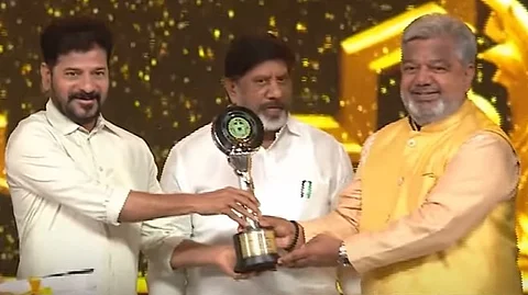 Three men are seen on stage during an award ceremony. The man on the left, wearing a white shirt, and the man on the right, dressed in a yellow kurta, are smiling as they hold a trophy together. A third man, also in a white shirt, stands in the middle behind them, observing the moment. The background is decorated with golden lights and patterns, giving a celebratory feel.