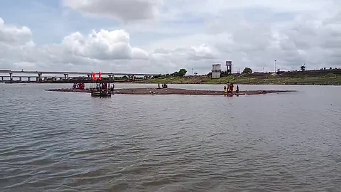 A group of people is gathered on a narrow sandbar in the middle of a river under a partly cloudy sky. A small wooden boat with red flags is anchored near the sandbar, and a concrete bridge and some structures are visible in the background on the riverbank. The scene suggests a ritual or local event taking place in a rural or semi-urban setting.