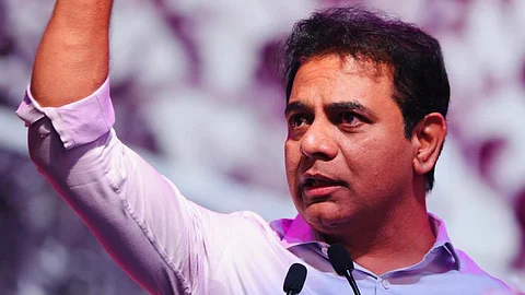 BRS leader KTR in a light purple shirt stands behind a podium adorned with pink flowers, raising his right fist in a powerful gesture. He speaks passionately into two microphones, with a blurred background of a crowd bathed in purple lighting, suggesting a public or political event.