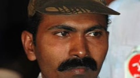 A man with a serious expression is seen wearing a camouflage-patterned cap with a red star emblem, symbolizing Maoist affiliation. He has a thick mustache and is dressed in a checkered shirt. The background is slightly blurred, showing another person wearing a badge with red and white colors.