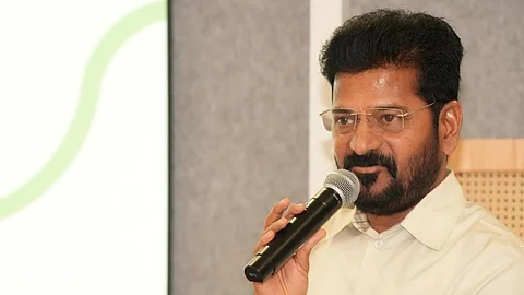 Telangana CM Revanth Reddy in a cream shirt is speaking into a microphone while standing at a podium. He has short black hair, a beard, and is wearing rimless glasses. A laptop is placed on the podium in front of him. The background features a grey panel and a projection screen with a green curved line.