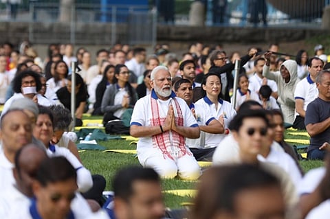 Prime Minister Narendra Modi sitting cross-legged with hands folded in a namaste gesture during a yoga session in a park, surrounded by participants dressed in white.