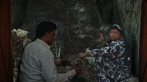 Two workers are seen manually stripping and sorting electrical wires inside a small room filled with coiled metal cables, likely as part of an informal recycling or scrap processing operation.
