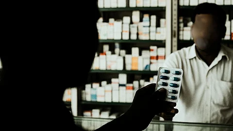 A man is holding up a strip of capsules. In front of him is a pharmacist and shelves stocked with medical drugs is seen behind the pharmacist.