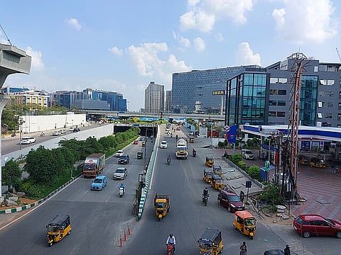 A bustling urban street scene in Gachibowli, Hyderabad, featuring multiple autorickshaws, cars, and motorbikes on a divided road. On the right side, there's a modern glass building with a petrol station beside it, while in the background, the large IKEA building stands prominently among other commercial high-rises. The sky is mostly clear with scattered clouds, and greenery lines parts of the road divider. A metro rail track is visible on the left.
