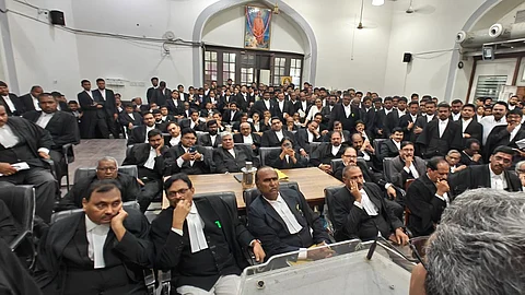 A large group of advocates, both men and women, dressed in black robes with white neckbands, are gathered in a courtroom or hall, seated and standing closely together. Most have serious or thoughtful expressions. The setting has arched doorways, a framed portrait on the wall, and air conditioning ducts visible above. A man with grey hair is seen from behind in the foreground, possibly addressing the group.