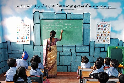 A teacher in a sari writes an English sentence on a chalkboard in a brightly painted classroom, as a group of uniformed primary school children sit facing her. The classroom walls are decorated with colourful educational posters and alphabet letters.