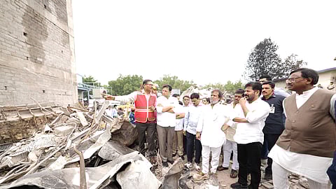CM Revanth Reddy with a group of government officials and emergency responders stand amid the rubble of a blast site, inspecting the extensive damage. One official wearing a red safety vest gestures toward the debris while speaking to others in the group, including men in white shirts, likely politicians or local leaders. The scene shows twisted metal, shattered concrete, and remnants of a collapsed structure, suggesting a recent explosion. The expressions of those present are serious and focused, indicating an on-site assessment of the destruction.