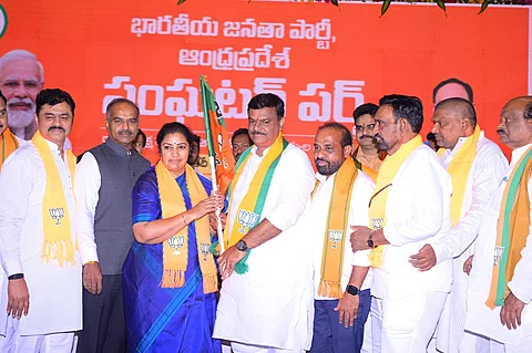 A group of Bharatiya Janata Party (BJP) leaders and members are seen on stage at a political event in Andhra Pradesh. A woman in a blue saree, wearing a BJP scarf, hands over a party flag to a man in white traditional attire, also wearing a BJP scarf. Other leaders stand alongside them, all dressed in white with orange BJP scarves, against a bright orange backdrop displaying the party's name in Telugu. Portraits of national BJP leaders, including Prime Minister Narendra Modi, are partially visible in the background. The atmosphere appears celebratory and symbolic of new membership or alliance.