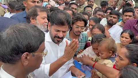 A large crowd gathers around CM Revanth Reddy in a white shirt. He is interacting with locals, including women and children, some of whom are holding documents and photographs. The expressions on the crowd’s faces range from hopeful to emotional, suggesting a response to a recent tragedy or crisis. Several people in the background are filming or taking photos, and security personnel are also visible.