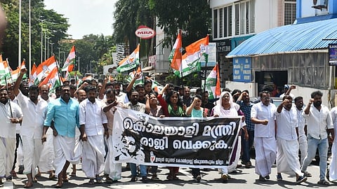 A large group of Youth Congress protestors march down a street in Thiruvananthapuram, holding a banner demanding the resignation of Health Minister Veena George. Many hold Congress flags and raise slogans, dressed in traditional Kerala attire.