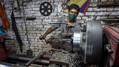 A man operates a lathe machine in a small workshop, wearing a striped shirt and a green face mask. Metal shavings are scattered around the machine, and the spinning metal component is in motion. The background features a white brick wall with hanging tools and machinery parts.