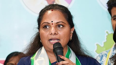 A woman in a navy blue saree is speaking into a microphone at a public event. She is wearing a white and green stole with "TELANGANA JAGRUTHI" and a Telangana state map logo printed on it. Her expression is serious and engaged, and she is gesturing with her hand while speaking. Other people are partially visible beside her.