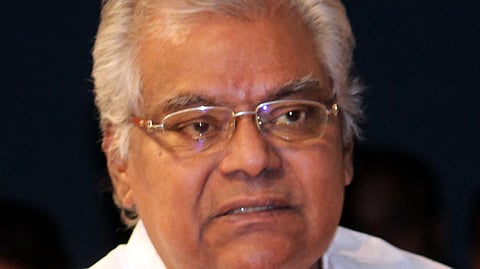 Elderly man with white hair and glasses wearing a white shirt, photographed indoors with a dark background.