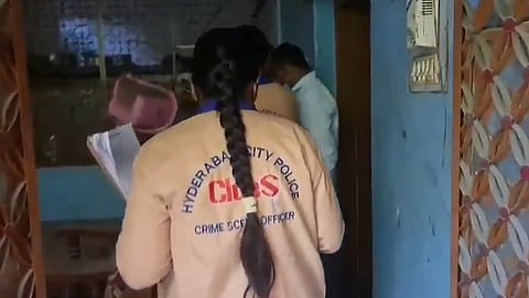 A female officer with a long braid, wearing a beige jacket labeled "Hyderabad City Police CLues Crime Scene Officer," is seen entering a room. She is holding documents or evidence sheets. Two other individuals, one also in uniform, are partially visible inside the building. The setting appears to be a modest residential or semi-commercial structure with blue walls and a patterned brown door.