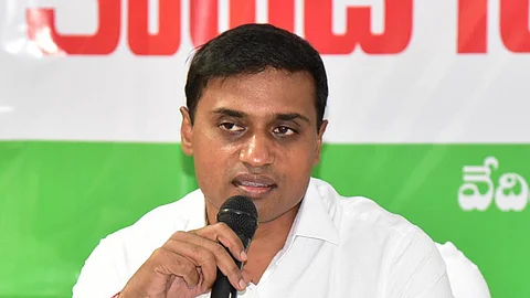 A man in a white shirt is speaking into a microphone at a press conference. He has short black hair and a serious expression. Behind him is a banner with Telugu text in red, white, and green colors.