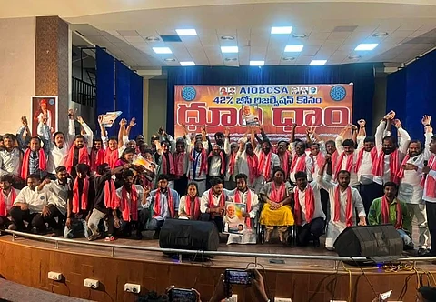 A large group of people, mostly men, pose on stage for a group photo at a public event. Many are wearing white shirts and red scarves, some holding up posters featuring political leaders. A banner in Telugu with the acronym "AIOBCSA" is displayed behind them, indicating the event is related to the demand for 42% reservation for OBCs. The atmosphere is energetic and united, with many individuals raising their fists in solidarity. A few people are seated at the front, including a woman in a wheelchair.