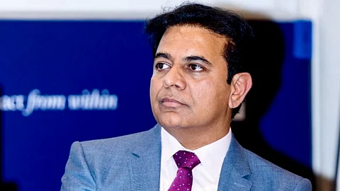 KTR in a blue suit with a maroon polka-dotted tie is seated on a chair, holding a microphone in his right hand. He has a serious expression and is looking slightly to his left. A clip-on microphone is attached to his lapel. The background is blue with indistinct text and logos.