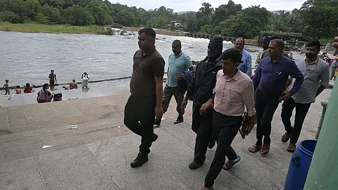Special Investigation Team officers walk with the whistleblower, whose face is covered, near the steps of the Netravathi Snana Ghatta in Dharmasthala as part of the probe into alleged secret burials.