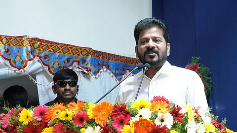 Revanth Reddy in a white shirt is speaking at a podium decorated with a vibrant arrangement of red, yellow, orange, and white flowers. He is flanked by two microphones. Behind him, a man in black sunglasses and a headset appears to be providing security. The background includes a blue curtain and a tent-like decorative canopy with colorful patterns.