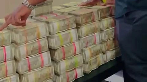 A man in a black shirt is standing beside a table stacked high with bundles of Indian currency notes, held together with colored bands. He appears to be pointing at one of the stacks, while resting his other hand on the money. The scene is indoors, with green and patterned curtains visible in the background.