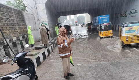 A traffic police officer in a raincoat stands on a flooded road under a bridge in Mettuguda, Hyderabad, Telangana, directing traffic during heavy rainfall. Water has accumulated heavily under the bridge, partially submerging the road and making it difficult for vehicles to pass. Several autorickshaws and a Swiggy Instamart delivery vehicle are seen navigating through the water. Other people in raincoats, including municipal workers, are present on the side of the road.