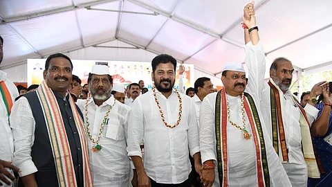 A group of Indian politicians, dressed mostly in white with tricolor garlands around their necks, stand together under a white tent at a political event. One man in the center raises another's hand in a gesture of unity. The atmosphere appears celebratory and formal, with several men wearing traditional Indian attire including Nehru caps. A backdrop behind them features political symbols and images.