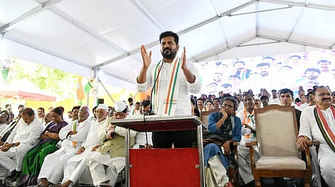 Image shows a man wearing a white shirt and an Indian National Congress tricolour scarf speaking at a podium under a large white tent. Several people, including men and women, are seated behind him, some wearing white attire and Gandhi caps. A large LED screen in the background displays an enlarged view of the scene. The atmosphere appears to be that of a political gathering or rally.