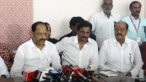 A group of men seated at a press conference table, with multiple microphones from various media outlets in front of them. Three men in white shirts are seated at the center, with others standing in the background, and a patterned wall partially visible on the left side.