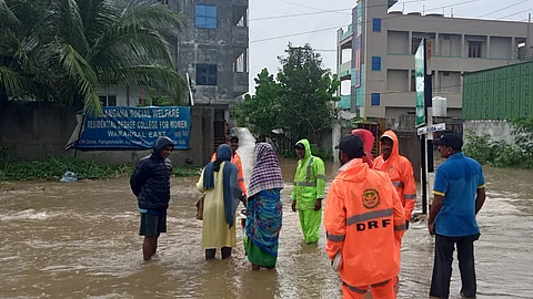 People, including municipal staff in orange and green raincoats marked “DRF,” stand in knee-deep floodwater on a street in Warangal, Telangana. Some residents are wearing shawls and rain jackets, while buildings and a sign for “Telangana Social Welfare Residential Degree College for Women, Warangal East” are visible in the background. The area is surrounded by trees and partially submerged roads.