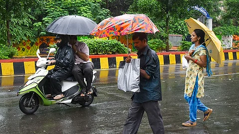 People walk and ride scooters on a wet road during rainfall. A man and woman on a scooter share a black umbrella, while another man walks with a colorful umbrella and shopping bags. A woman in traditional attire walks with a yellow umbrella. Trees and a painted roadside wall are visible in the background.
