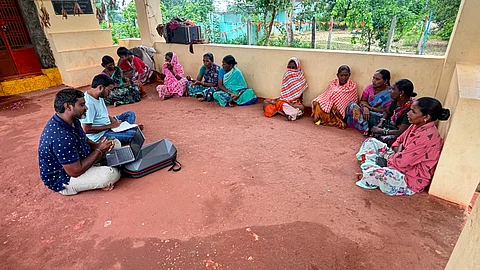 A group of Chenchu tribal women in Telangana sit together in a semi-circle on the floor of a shaded outdoor veranda. They are dressed in colourful sarees and shawls, some with patterned headscarves. Two men, seated cross-legged in front of them, are using a laptop and notebook, appearing to record or discuss something with the women. The background shows greenery, trees, and a compound wall, suggesting a rural setting. The scene reflects a community meeting.