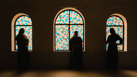 Three nuns in dark habits stand silhouetted in front of colourful stained glass windows inside a church, with the vibrant blue, orange, and white glass creating a striking contrast against their shadowed figures.