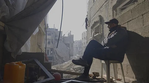 A man sits on a plastic chair in the shadow of a bombed-out building in Gaza, looking out at rubble and destroyed homes, in a still from the Palestinian anthology From Ground Zero.