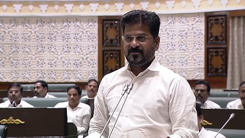 A man in a white shirt, wearing glasses, is speaking at a podium with microphones in a legislative assembly hall. Several men in white shirts are seated behind him, listening attentively. The background has patterned wall panels and wooden designs.