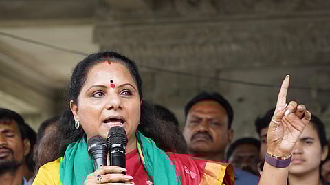 A woman wearing a red saree with a yellow blouse and a green scarf speaks passionately into two microphones while gesturing with her right hand raised. She has a red bindi on her forehead and is surrounded by people in the background.