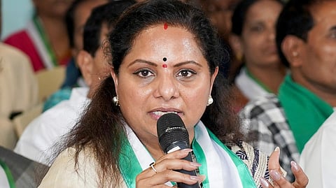 A woman wearing a cream and blue saree with a green and white sash is speaking into a microphone at an event. She has a red bindi on her forehead, pearl earrings, and is gesturing with her hand while addressing the audience. Several people are seated in the background, listening attentively.